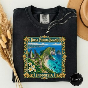 Könnte beinhalten: Ein schwarzes T-Shirt mit Rundhalsausschnitt und einem Grafikdruck der Insel Nusa Penida, Indonesien, eingerahmt von einer goldenen Blumenbordüre. Das Design zeigt eine Küstenklippe, blaues Meer, Mantarochen und weiße Plumeria-Blüten.