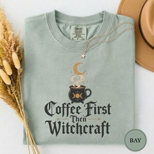 Può includere: Una t-shirt a maniche corte color verde salvia con la grafica di un calderone nero da cui sale vapore verso una luna crescente. Il testo recita Coffee First Then Witchcraft. Una delicata collana a catena argentata con una stellina poggia sul colletto.