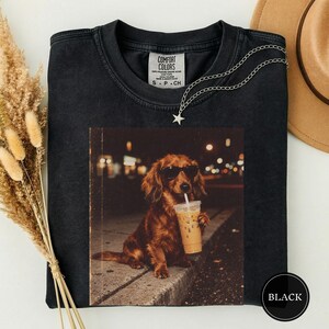 May include: A black crewneck t-shirt featuring a graphic print of a long-haired brown dog wearing black sunglasses and holding a plastic cup with a straw. The shirt has a Comfort Colors label inside the collar and a silver star pendant necklace.