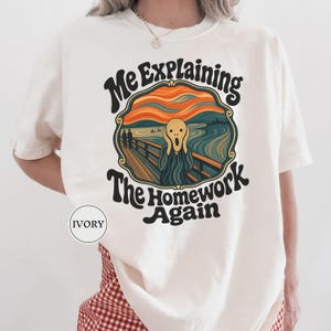 May include: An ivory short-sleeve t-shirt featuring a graphic print of a parody of The Scream painting. The text reads Me Explaining The Homework Again in a dark, wavy font. The graphic includes orange, blue, and black colors.