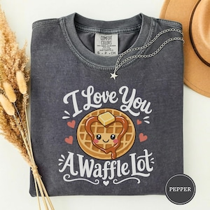 May include: A charcoal grey t-shirt featuring a graphic of a smiling waffle with a heart-shaped syrup drizzle and a pat of butter. The text reads I Love You A Waffle Lot. The shirt has a crew neckline and a Comfort Colors brand label at the collar.