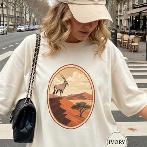 Peut inclure: Un t-shirt ivoire avec un motif ovale central représentant un oryx debout sur une falaise désertique surplombant un paysage de dunes de sable et un arbre isolé sous un ciel nuageux. Le t-shirt est porté avec un sac noir à bandoulière chaîne.