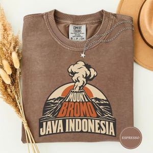 Peut inclure: Un t-shirt marron à manches courtes orné d'un motif de volcan en éruption avec le texte MOUNT BROMO JAVA INDONESIA. Le vêtement présente un col rond et un collier à chaîne argentée avec un petit pendentif en forme d'étoile sur le col.