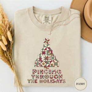May include: An ivory crewneck t-shirt featuring a graphic of a Christmas tree constructed from various geometric quilt-style patterns in shades of red, green, and tan. Below the tree, the text reads PIECING THROUGH THE HOLIDAYS in a matching font.