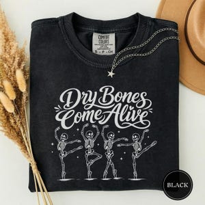 May include: A black crewneck t-shirt featuring a white graphic of four dancing skeletons and the text "Dry Bones Come Alive" in a cursive script. The shirt has a Comfort Colors label at the inner neckline and a silver star-shaped pendant necklace.