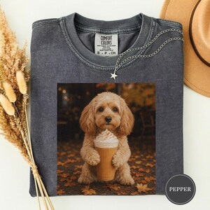 Könnte beinhalten: Ein anthrazitfarbenes T-Shirt mit Rundhalsausschnitt und einem Grafikdruck eines hellbraunen Hundes, der einen beigen Kaffeebecher mit Schlagsahne hält. Das Shirt hat eine Vintage-Waschung und ein Etikett mit der Aufschrift COMFORT COLORS. Eine silberne Sternkette.