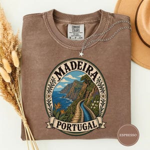 Puede incluir: Una camiseta de manga corta marrón con un estampado gráfico de un paisaje costero de Madeira, Portugal. El diseño incluye un camino sinuoso, acantilados, el océano y flores dentro de un marco ovalado con el texto Madeira Portugal.