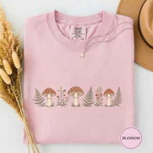 May include: A light pink short-sleeve t-shirt featuring a graphic print of three brown mushrooms interspersed with green ferns and small wildflowers. The inner neck label reads COMFORT COLORS S P CH. A silver star-shaped necklace rests on the fabric.
