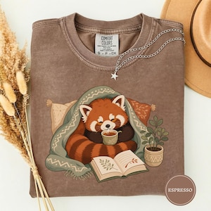 Puede incluir: Una camiseta marrón de cuello redondo con un gráfico de un panda rojo envuelto en una manta, sosteniendo una taza y leyendo un libro abierto. La camiseta tiene una etiqueta Comfort Colors en el cuello y se combina con un collar de estrella plateado y un sombrero beige.