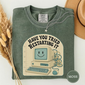 Puede incluir: Una camiseta de cuello redondo color verde musgo con un gráfico de una computadora antigua con una cara sonriente en la pantalla. El texto dice HAVE YOU TRIED RESTARTING IT. Un collar con dije de estrella plateada descansa en el cuello.
