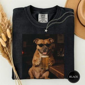May include: A black t-shirt featuring a graphic of a brown dog wearing black sunglasses and holding a glass mug of beer. The shirt has a Comfort Colors label at the neck and a silver-toned chain necklace with a star pendant resting on the fabric.