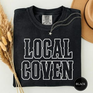 May include: A black crewneck t-shirt featuring the white text LOCAL COVEN in a bold, collegiate-style font. The shirt has a Comfort Colors label at the inner neckline and is accented by a delicate silver-toned chain necklace with a small star charm.
