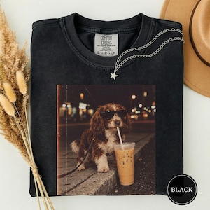 Può includere: Una t-shirt nera a girocollo con una stampa grafica di un cane marrone e bianco che indossa occhiali da sole neri, in piedi accanto a un caffè freddo con cannuccia. La maglietta ha un'etichetta Comfort Colors sul collo e una collana a stella.
