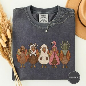 Könnte beinhalten: Ein anthrazitfarbenes T-Shirt mit Rundhalsausschnitt und einer Grafik von fünf Truthähnen in verschiedenen Kostümen, darunter ein Schnurrbart, Kuhflecken, Schafwolle, ein Flamingo und eine Topfpflanze. Mit Comfort Colors Etikett und Sternkette.