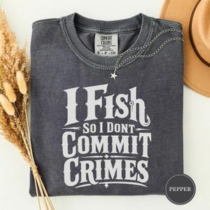 Pode incluir: Uma t-shirt de manga curta cinzento-carvão com acabamento de lavagem vintage. A frente apresenta um texto branco que diz I Fish So I Dont Commit Crimes. Um colar prateado com um pequeno pendente de estrela repousa sobre o decote da camisa.