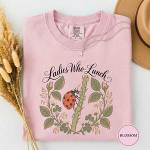 Puede incluir: Una camiseta rosa claro con un gráfico de una mariquita roja sobre un tallo verde con espinas, rodeada de hojas verdes y flores de trébol blancas. El texto "Ladies Who Lunch" aparece en negro sobre la ilustración. Un collar de estrella plateado.