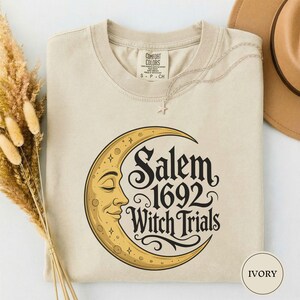 May include: An ivory short-sleeve t-shirt featuring a graphic of a yellow crescent moon with a face. The text reads Salem 1692 Witch Trials in a black decorative font. The shirt has a crew neckline and a Comfort Colors brand label at the neck.