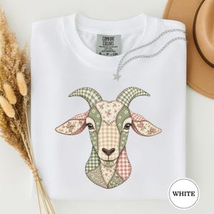 May include: A white short-sleeve t-shirt featuring a graphic of a goat head composed of various patchwork patterns in shades of green, pink, and beige. The shirt has a Comfort Colors label at the neck and a silver-toned star charm necklace.