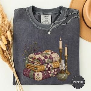 Könnte beinhalten: Ein anthrazitfarbenes T-Shirt mit Rundhalsausschnitt und einem Grafikdruck, der einen Stapel aus drei gefalteten Patchwork-Decken, eine kleine Vase mit Trockenblumen, zwei hohe Kerzenständer mit brennenden Kerzen und eine kleine Öllampe zeigt.