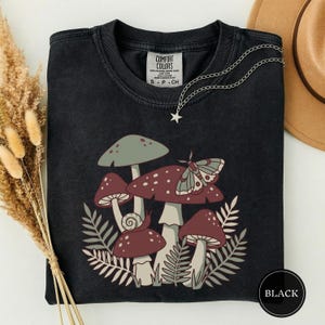 May include: A black crewneck t-shirt featuring a graphic print of various mushrooms, a moth, and a snail nestled among fern leaves. The shirt has a Comfort Colors label at the inner neckline and a silver-toned star charm necklace resting on top.