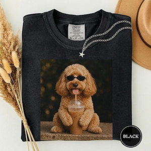 May include: A black crewneck t-shirt featuring a graphic print of a golden-brown poodle wearing black sunglasses and holding a clear plastic cup with a straw. The shirt has a Comfort Colors label at the inner neckline and a silver star necklace.