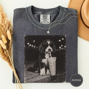 May include: A charcoal gray crew neck t-shirt featuring a graphic print of a fluffy dog wearing sunglasses and drinking from a cup with a straw. The shirt has a Comfort Colors label at the inner neckline and a small star charm necklace resting on it.