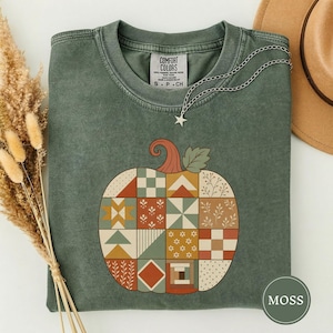 May include: A moss green crewneck t-shirt featuring a large pumpkin graphic composed of various geometric and floral quilt-like patterns in shades of orange, cream, and sage green. The shirt has a Comfort Colors label at the inner neckline.