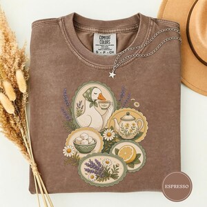 Puede incluir: Una camiseta marrón de cuello redondo con un estampado gráfico de un ganso bebiendo té, una tetera floral, un cuenco de terrones de azúcar, una rodaja de limón y ramas de lavanda. La prenda tiene una etiqueta Comfort Colors y un collar de estrella.