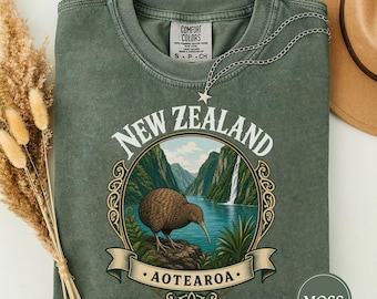 May include: A moss green crewneck t-shirt featuring a graphic print of a kiwi bird standing on a rock before a mountain landscape with a waterfall. The text reads NEW ZEALAND and AOTEAROA. A silver star pendant necklace rests on the neckline.