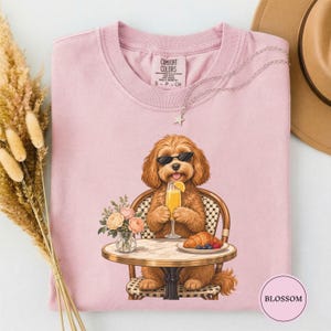 May include: A light pink t-shirt featuring a graphic of a brown dog wearing black sunglasses, sitting at a round table. The dog holds a glass of orange juice next to a plate with a croissant and berries. A small vase of flowers sits on the table.