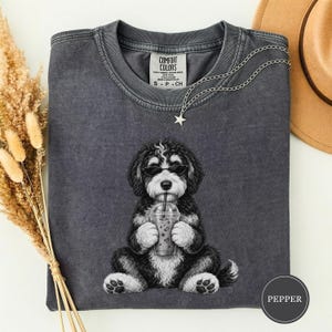 May include: A charcoal gray crewneck t-shirt featuring a graphic of a black and white dog wearing sunglasses and holding a plastic cup with a straw. The shirt has a Comfort Colors label at the neck and a small star charm on a silver chain.