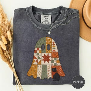Puede incluir: Una camiseta de cuello redondo gris carbón con un gráfico de un fantasma hecho de patrones de patchwork en tonos apagados naranja, verde y crema. Un pequeño gato negro con un pañuelo rojo se sienta en la base del fantasma. La camiseta tiene una etiqueta Comfort Colors.