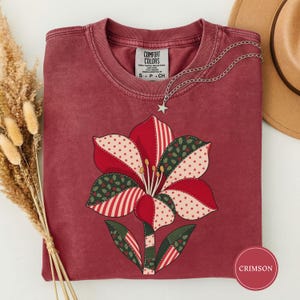 May include: A crimson crewneck t-shirt featuring a large, stylised flower graphic composed of patchwork-style segments in red, white, and dark green with star and holly leaf patterns. The shirt has a tag labelled Comfort Colors and a star charm necklace.
