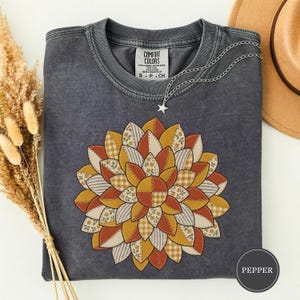 May include: A charcoal grey short-sleeve t-shirt featuring a large, circular floral graphic composed of patchwork-style petals in shades of mustard yellow, burnt orange, cream, and white, with various patterns including gingham and small floral prints.