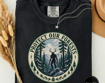 May include: A black crewneck t-shirt featuring a circular graphic with the text "PROTECT OUR FORESTS" surrounding an illustration of a silhouetted creature with antlers standing on a rocky outcrop in a forest with mountains in the background.