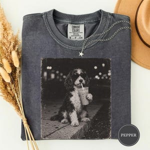 Puede incluir: Una camiseta de manga corta gris carbón con un gráfico en blanco y negro de un perro con gafas de sol que sostiene un vaso de plástico con pajita. La camiseta tiene cuello redondo y una etiqueta Comfort Colors impresa en el interior.
