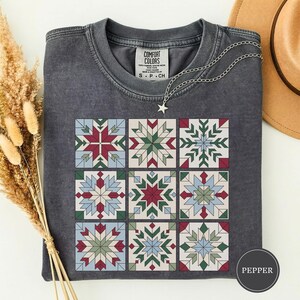 May include: A charcoal gray short-sleeve t-shirt featuring a grid of nine square quilt-style blocks with geometric star patterns in shades of red, green, and light blue. The shirt has a crew neckline and a Comfort Colors brand label at the neck.
