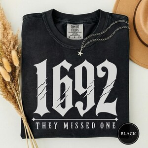 May include: A black short-sleeved t-shirt featuring the white text 1692 and the phrase THEY MISSED ONE. The numbers have a distressed claw mark design. A silver-toned chain necklace with a small star pendant rests on the neckline of the shirt.