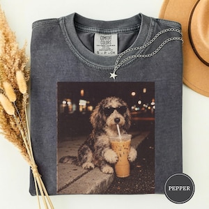 Op de afbeelding: Een antracietgrijs t-shirt met ronde hals en een grafische print van een pluizige hond met een zwarte zonnebril die een plastic beker met een rietje vasthoudt. Het shirt heeft een Comfort Colors-label en een zilverkleurige sterrenketting.