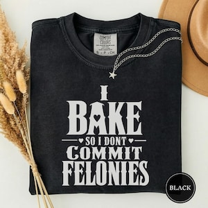 May include: A black short-sleeved t-shirt with white text that reads I BAKE SO I DONT COMMIT FELONIES. The shirt features a Comfort Colors brand label at the inner neckline and a small silver-toned star charm necklace resting on the fabric.