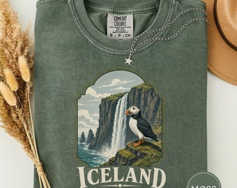 Op de afbeelding: Een mosgroen T-shirt met ronde hals en een afbeelding van een papegaaiduiker op een klif bij een waterval. De tekst luidt ICELAND LAND OF ICE. Een zilveren ketting met sterhanger ligt op de halslijn. Het shirt heeft een Comfort Colors label.