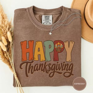 Puede incluir: Una camiseta de cuello redondo marrón con efecto desgastado, que presenta el texto HAPPY THANKSGIVING en letras grandes y coloridas. La letra P contiene un pequeño gráfico de un pavo. Un collar con colgante de estrella plateada descansa en el cuello.