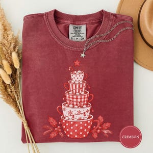 May include: A crimson crewneck t-shirt featuring a graphic of five stacked mugs forming a Christmas tree shape. The mugs have various red and white patterns including polka dots, stripes, and snowflakes. A small star sits atop the stack.