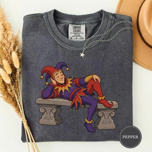 Puede incluir: Una camiseta de manga corta gris carbón con un gráfico de un bufón con traje morado y rojo recostado en un banco de piedra. La camiseta tiene cuello redondo y una etiqueta Comfort Colors. Un collar con colgante de estrella plateado descansa sobre la tela.