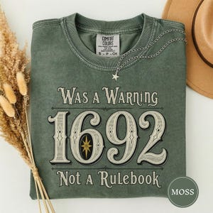 Könnte beinhalten: Ein moosgrünes Kurzarm-T-Shirt mit einem Grafikdruck im Vintage-Stil. Der Text "Was a Warning 1692 Not a Rulebook" ist in einer dekorativen Schriftart gehalten und enthält ein zentrales Sternsymbol. Das Shirt hat einen Rundhalsausschnitt und ein Comfort Colors-Etikett.