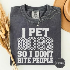 Könnte beinhalten: Ein anthrazitfarbenes Kurzarm-T-Shirt mit Vintage-Waschung. Auf der Vorderseite befindet sich ein weißer Schriftzug I PET DOGS SO I DONT BITE PEOPLE, wobei das Wort DOGS in einem Schachbrettmuster gestaltet ist. Eine silberne Stern-Halskette liegt bei.