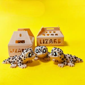 May include: Three articulated lizard figurines with tan bodies and dark brown spots, each featuring large white eyes. Behind the figurines are two light tan plastic carrying cases, each with a handle and the word LIZARD printed on the side.