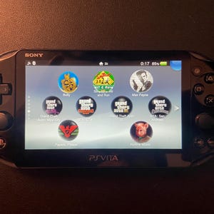 Può includere: Una console di gioco portatile Sony PlayStation Vita nera con uno schermo lucido che mostra un menu di icone di gioco, tra cui Bully, The Simpsons Hit and Run, Max Payne, titoli Grand Theft Auto, Papers Please e Hotline Miami.