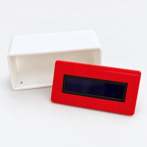 Puede incluir: Una carcasa rectangular de plástico blanco con la parte frontal abierta y un pequeño orificio en la superficie interior inferior. Una placa frontal rectangular roja separada presenta una pantalla digital azul oscuro con borde negro.