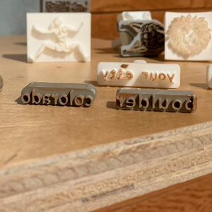 May include: A collection of clay stamps and a plastic container. The stamps feature text including 2026, colorado, boulder, and your state. One large oval black stamp displays 2026 and colorado. Several smaller white and metallic rectangular stamps are present.